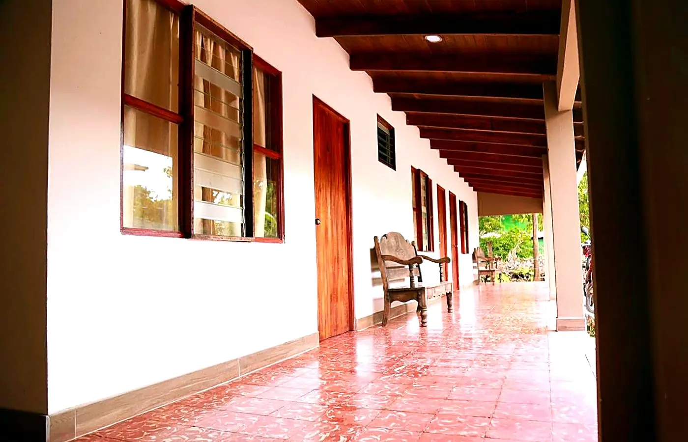 Corredor del hotel con piso de baldosa de barro, paredes blancas, puertas de madera oscura y mecedoras